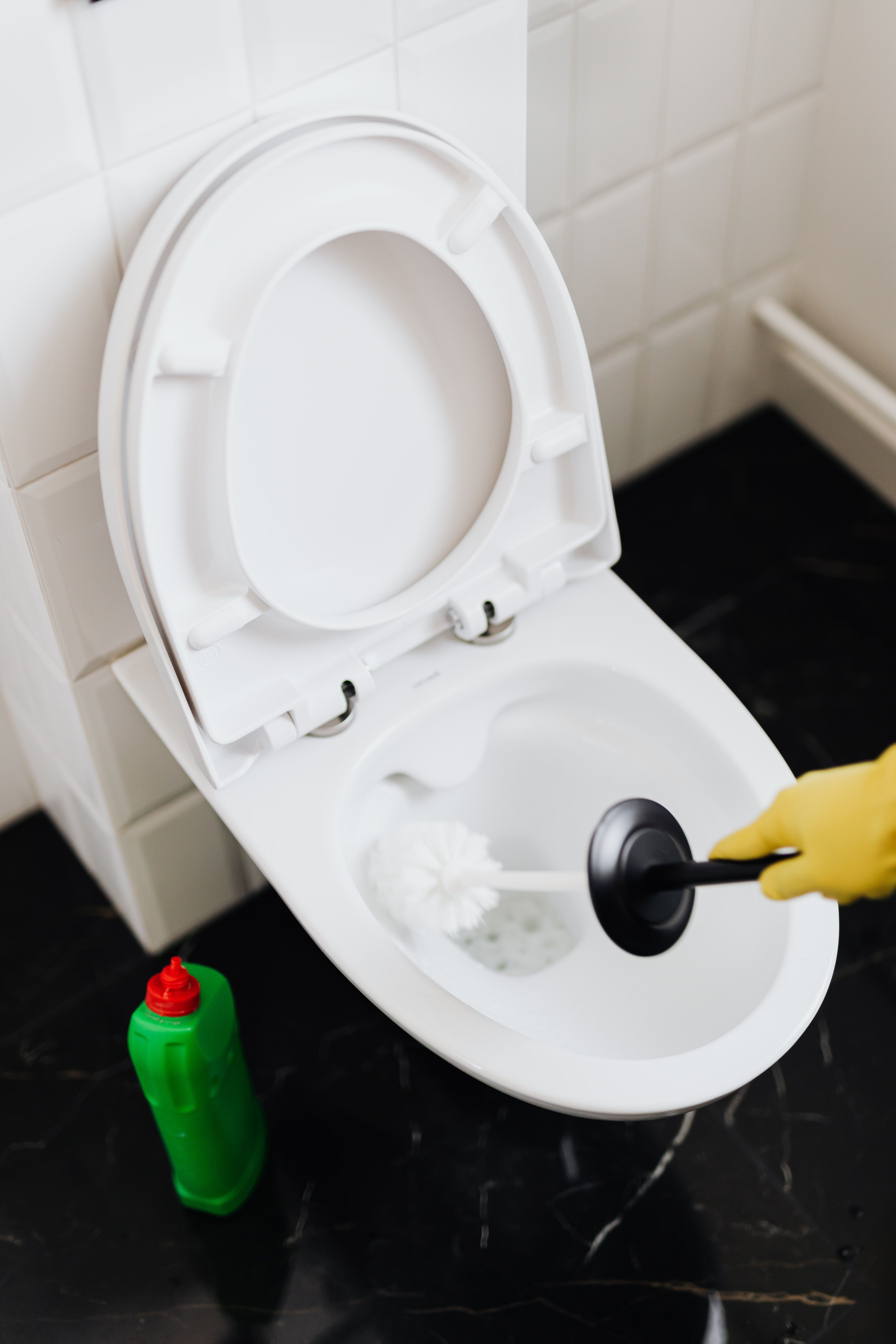 A close-up view of a hand in a yellow rubber glove using a white brush to scrub a clean toilet bowl, with a green cleaning bottle sitting on a black marble floor.